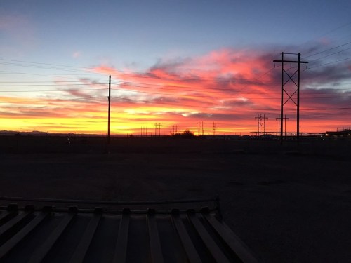 Vibrant sunset with pink and orange hues over a landscape with power lines and a silhouette of distant mountains.