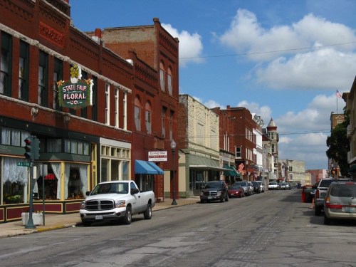A quaint street lined with historic buildings, shops, and parked cars under a partly cloudy sky.