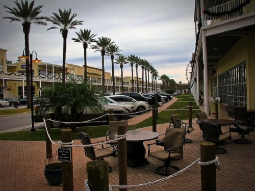 A palm-lined walkway with parked cars and outdoor seating, set against a cloudy sky.