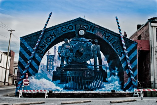 Mural of Union Cotton Mills featuring a steam train and two workers, set against a cloudy sky.