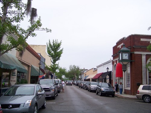 A quiet street lined with shops and parked cars, featuring a clock tower and overcast sky.