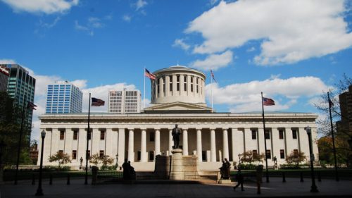 The Ohio Statehouse, a grand building with a dome, surrounded by flags and a clear blue sky.