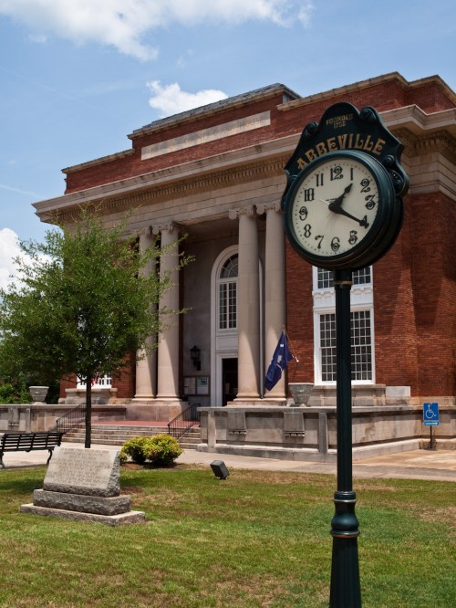 A vintage clock stands in front of a large brick building with columns, surrounded by green grass and trees.