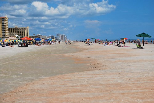 A sunny beach scene with people relaxing under umbrellas, buildings in the background, and a clear blue sky.
