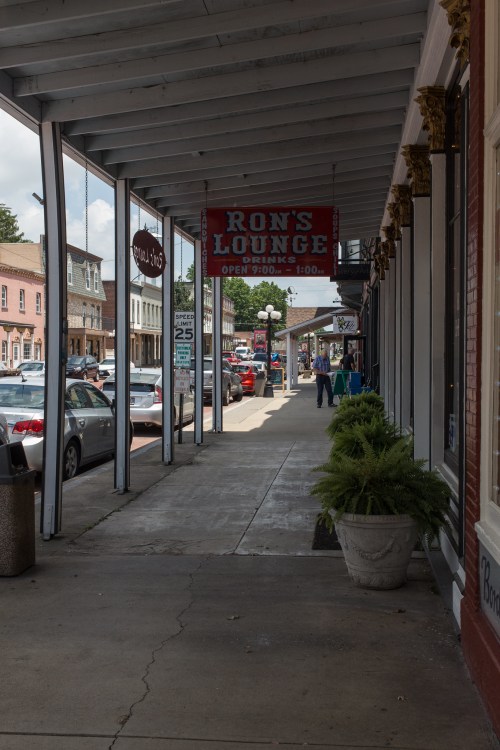 A street view featuring a sign for Ron's Lounge, with parked cars and shops under a covered walkway.