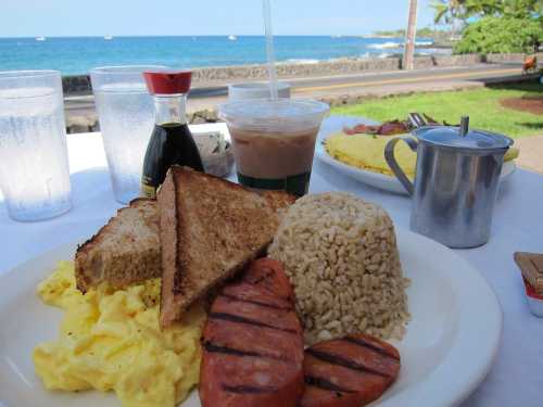 A plate of scrambled eggs, grilled sausage, rice, and toast with drinks and a scenic ocean view in the background.