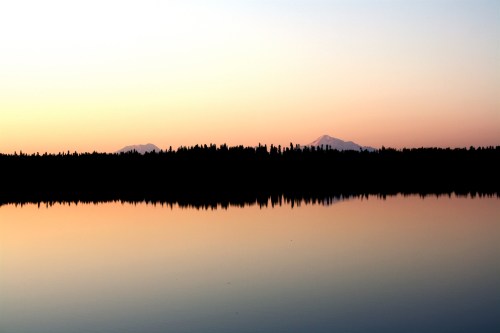 A serene lake at sunset, reflecting mountains and a colorful sky, framed by dark silhouettes of trees.