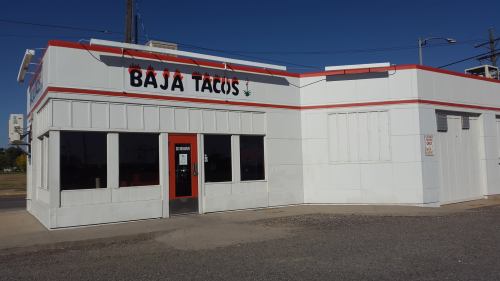 Exterior of a Baja Tacos restaurant, featuring a white building with red accents and large windows. Clear blue sky above.