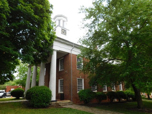 Historic brick building with white columns and a clock tower, surrounded by green trees and shrubs.