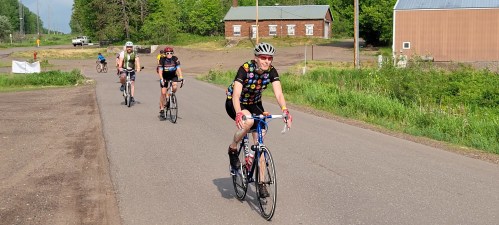 A group of cyclists riding on a rural road, surrounded by greenery and buildings in the background.