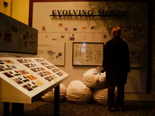 A person stands in front of an exhibit titled "Evolving Money," surrounded by large bags and display panels.