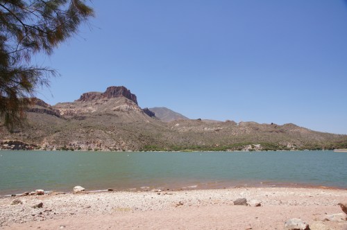 A serene lakeside view with a sandy shore, mountains in the background, and clear blue skies.