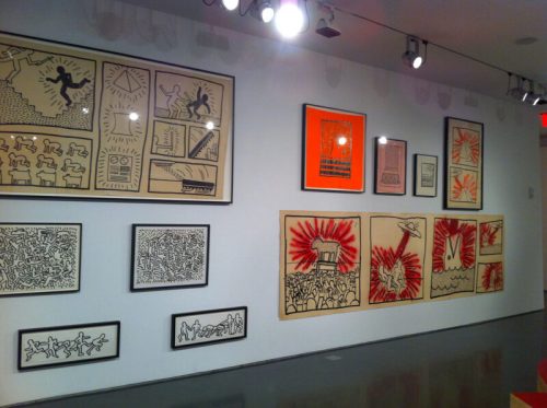 A gallery wall displaying framed artworks in black and red, featuring abstract figures and dynamic patterns.