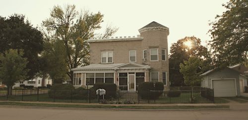 A large, historic house with a turret, surrounded by greenery and a fence, illuminated by a sunset in the background.
