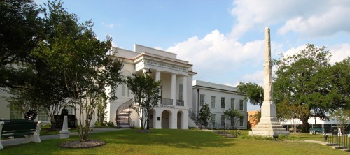 Historic building with columns, surrounded by trees and grass, featuring a tall monument in the foreground.
