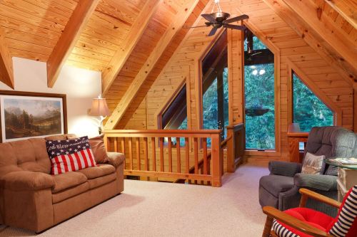 Cozy wooden loft with sloped ceilings, a sofa, armchair, and large windows overlooking a forested view.