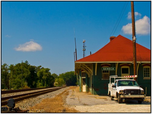 A vintage train station named "Seneca" beside railway tracks, with a white truck parked nearby under a clear blue sky.