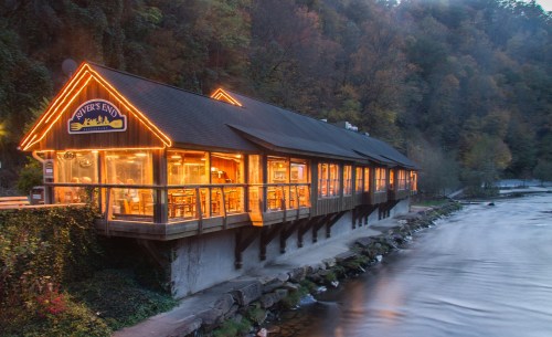 A riverside restaurant with warm lights, nestled among trees, reflecting on the water at dusk.