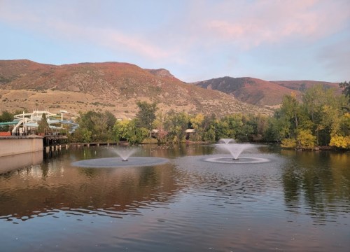 A serene lake with two fountains, surrounded by trees and mountains under a pastel sky at sunset.
