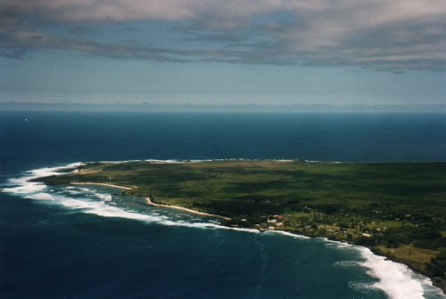Aerial view of a coastal landscape with green land, waves crashing on the shore, and a cloudy sky above.