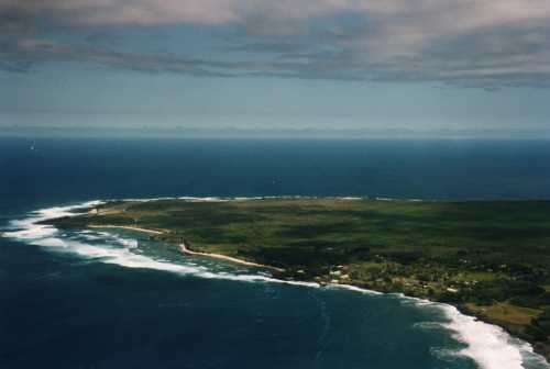 Aerial view of a coastal landscape with green land, waves crashing on the shore, and a cloudy sky above.