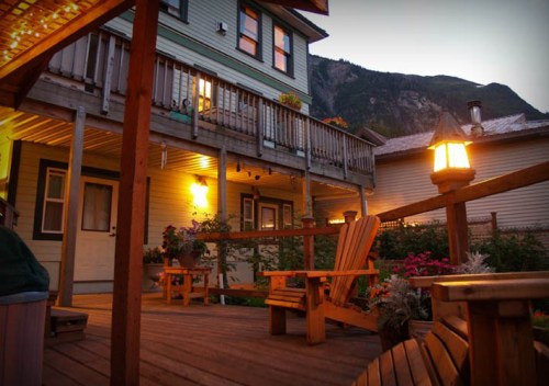 A cozy wooden deck with chairs, surrounded by flowers and a mountain backdrop at dusk. Warm lights illuminate the scene.