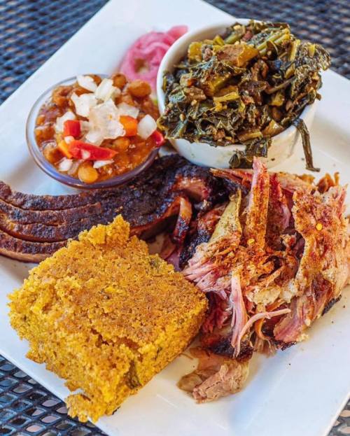 A plate of barbecue featuring pulled pork, brisket, cornbread, collard greens, and baked beans.