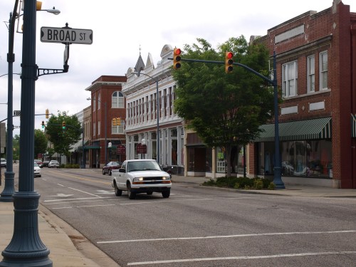 A quiet street scene with shops, traffic lights, and a white car driving on Broad Street. Overcast sky above.