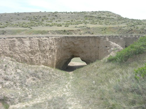 A natural archway formed in a sandy landscape, surrounded by grass and hills under a cloudy sky.