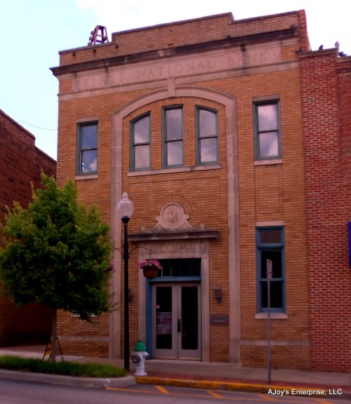 Historic brick building with "First National Bank" engraved above the entrance, featuring large windows and a tree nearby.