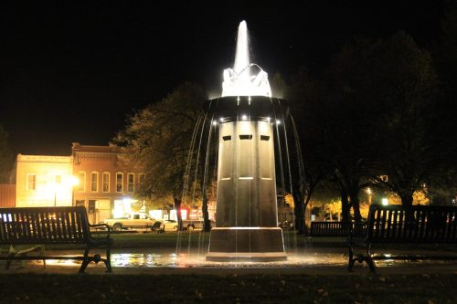 A lit fountain at night, surrounded by benches and trees, with a charming town in the background.