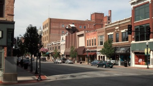 A quiet downtown street lined with shops and trees, featuring brick buildings and parked cars under a clear sky.