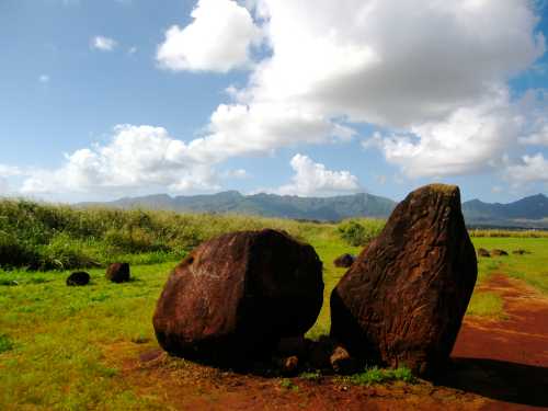 Two large, weathered rocks sit on green grass under a blue sky with fluffy clouds and distant mountains in the background.