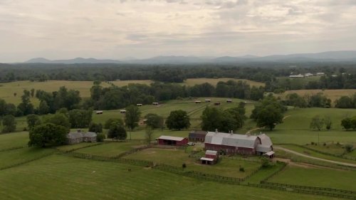 Aerial view of a rural farm with red barns, green fields, and distant mountains under a cloudy sky.