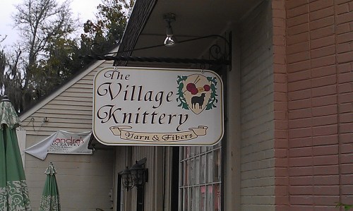 Sign for "The Village Knittery" featuring yarn and fibers, hanging from a building with decorative elements.