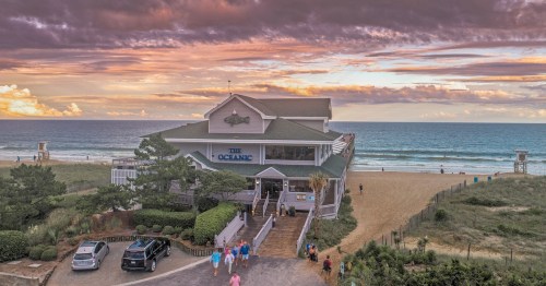 A beachside restaurant, "The Oceanic," with a colorful sunset and people walking on the sandy shore.