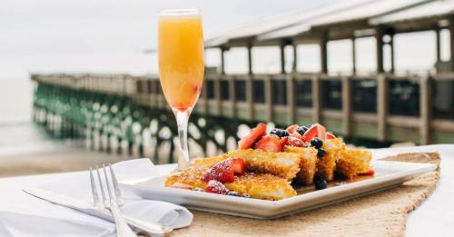 A plate of French toast topped with berries, alongside a mimosa, with a beach pier in the background.