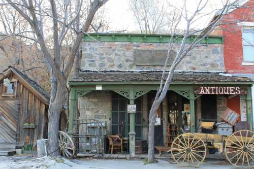 A rustic antique shop with a stone facade, wooden signage, and vintage carts outside, surrounded by bare trees.