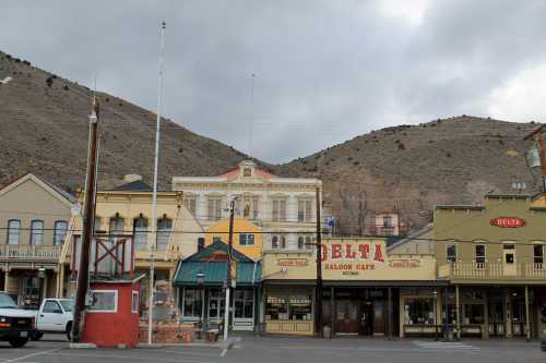 Historic buildings line a street in a small town, with mountains in the background under a cloudy sky.