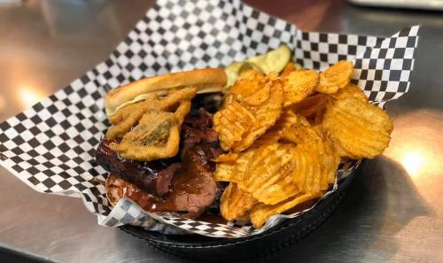 A basket of barbecue sandwich with pickles and crispy potato chips on a checkered paper.