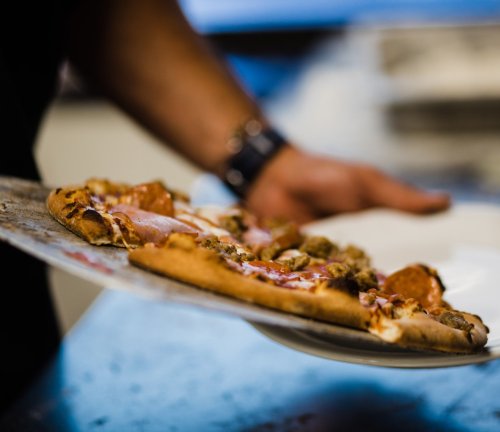 A person holding a pizza on a metal tray, with toppings and sauce visible, ready to be served.