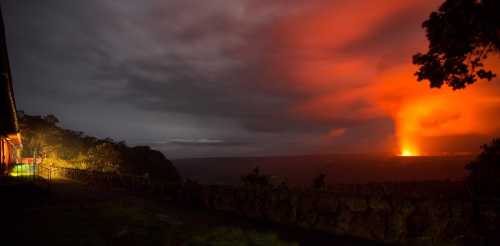 A dramatic night scene of a volcanic eruption illuminating the sky, with a house and trees in the foreground.