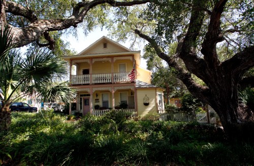 A charming yellow two-story house surrounded by lush greenery and large trees, featuring a front porch and an American flag.