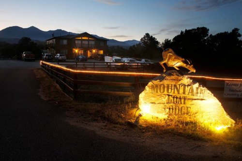 A stone sign for Mountain Goat Lodge illuminated at dusk, with a lodge and mountains in the background.