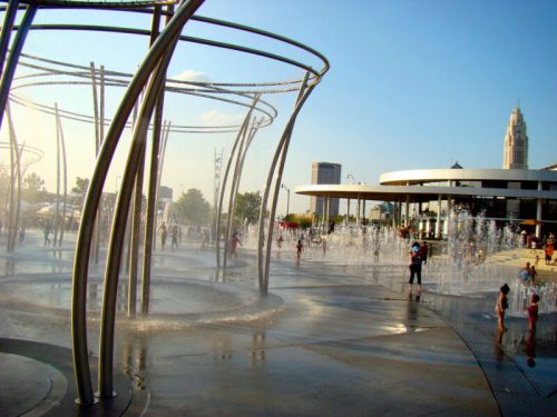 A public fountain with water jets, children playing, and a city skyline in the background on a sunny day.