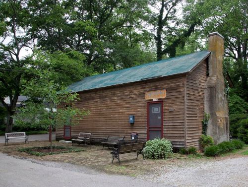 A rustic wooden building with a green roof, surrounded by trees and benches, set in a peaceful outdoor area.