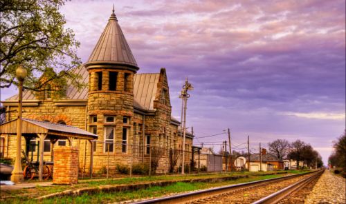 Historic stone train station beside railway tracks at sunset, with a colorful sky and surrounding trees.