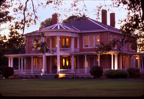 A large, elegant two-story house with a wraparound porch, illuminated at dusk, surrounded by lush greenery.