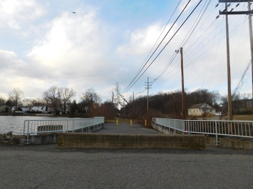 A concrete dock by a river, with power lines overhead and trees in the background under a cloudy sky.