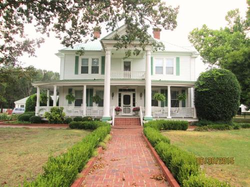 A large, two-story white house with green shutters, a front porch, and a brick pathway surrounded by greenery.